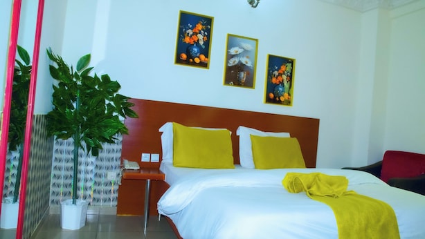 Une chambre d’hôtel avec un lit, deux oreillers jaunes, une table de chevet, un téléphone, une chaise rouge et trois tableaux représentant des motifs floraux, encadrés et accrochés au mur.