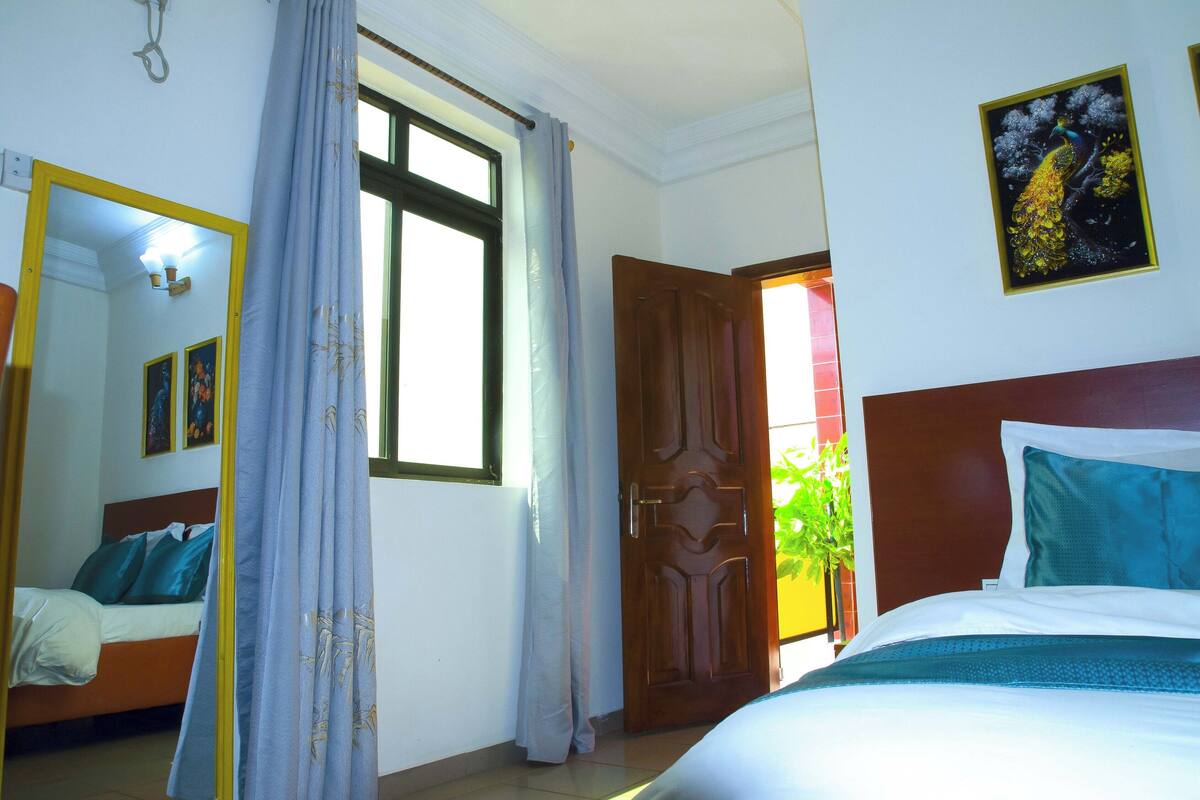 A hotel room with a bed, a window with curtains, a door, and a wall-mounted picture.