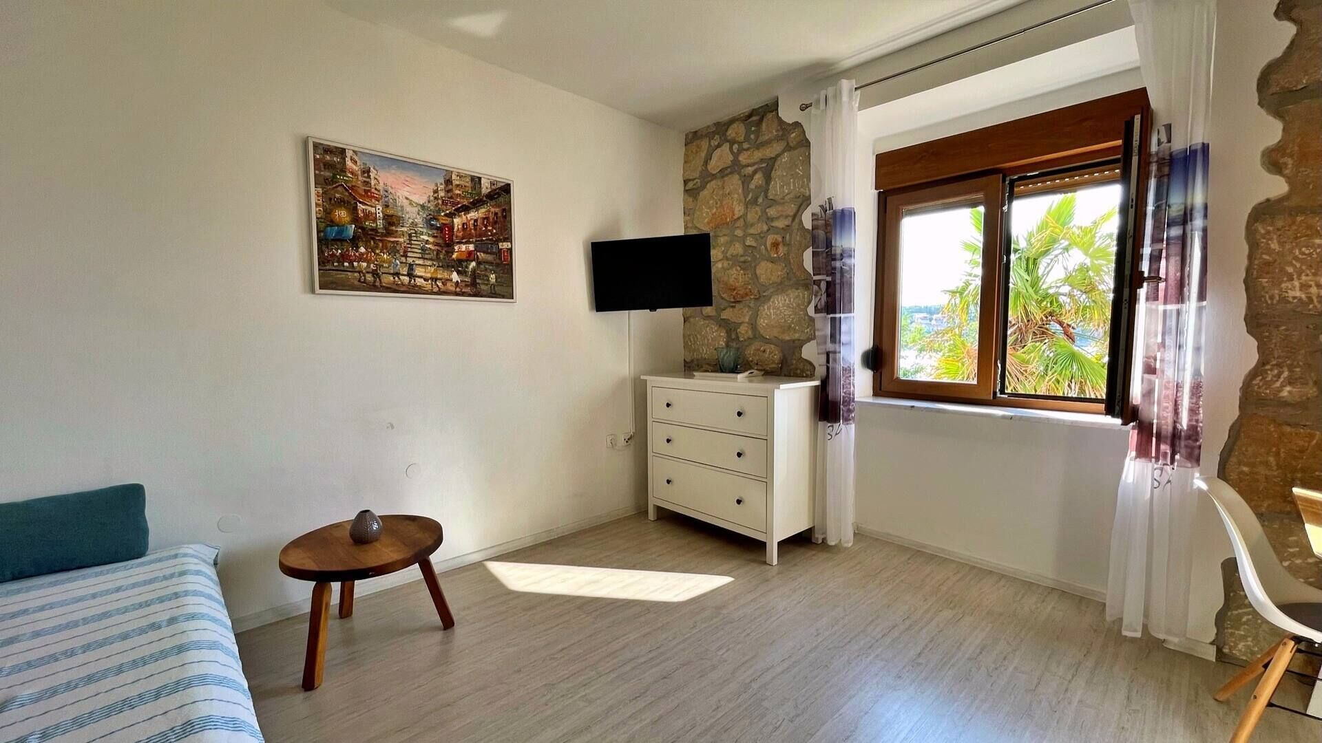 Une chambre avec un lit, une petite table, une commode, un téléviseur et une fenêtre donnant sur un paysage verdoyant.