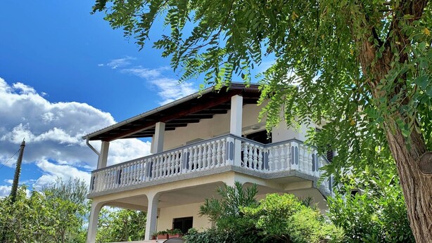 Una casa de dos pisos con balcón, rodeada de follaje y un cielo despejado.