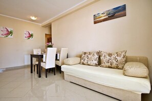 A living room with a sofa, two floral cushions, a dining table with chairs, and a painting on the wall.