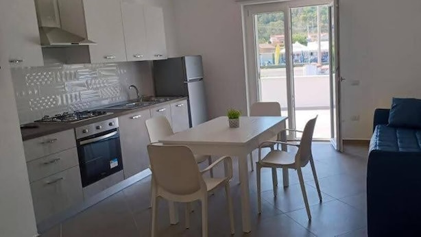 A modern kitchen with white cabinets, a dining table with chairs, and a dark blue sofa in a bright room with a large window. - Tenuta Nonna Peppa Luxury apartment (Centola)