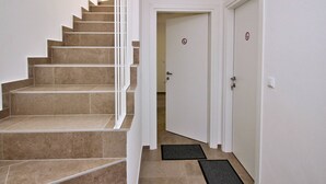 Un escalier aux marches recouvertes de carreaux beiges et doté de rampes blanches, menant à une porte surmontée d’un paillasson.