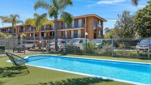A swimming pool with a chair, surrounded by a fenced area with parked cars and a building with multiple balconies. - Coastal Comfort in Batehaven (Batehaven)