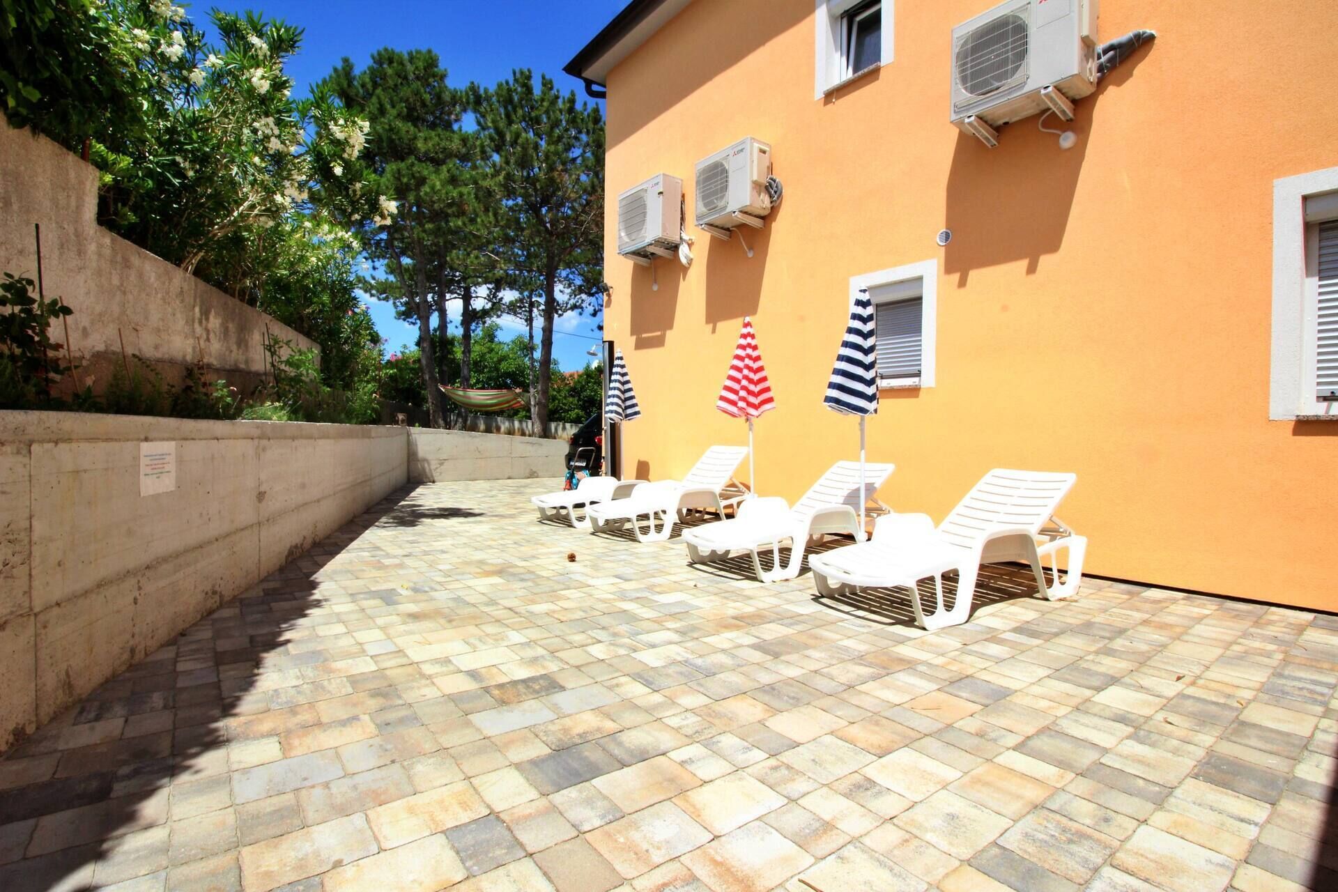 A sunlit outdoor seating area with white lounge chairs and striped umbrellas, a concrete wall, and air conditioning units on the building.