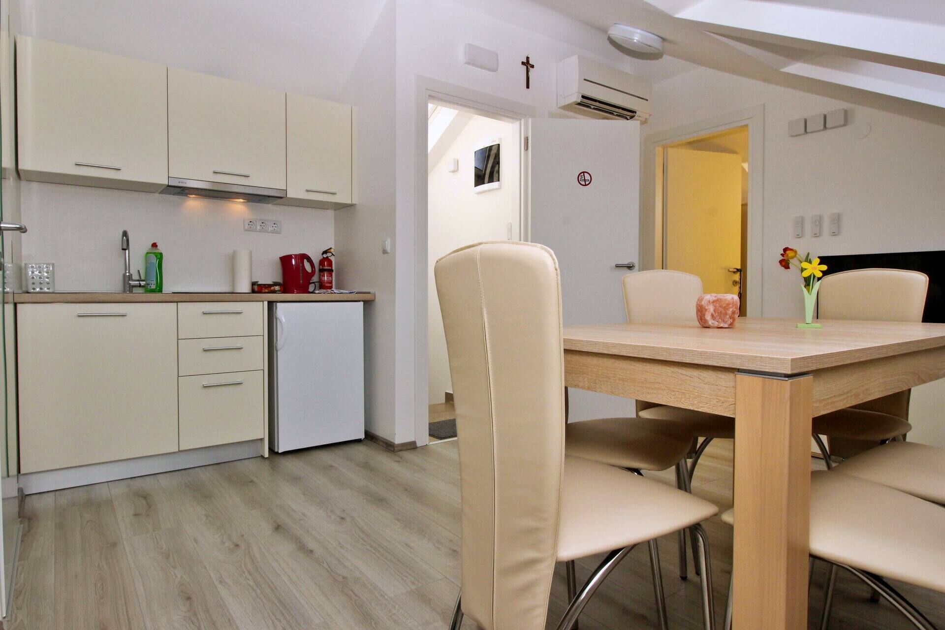 A modern kitchen with white cabinets, a small refrigerator, and a dining area with a wooden table and beige chairs.