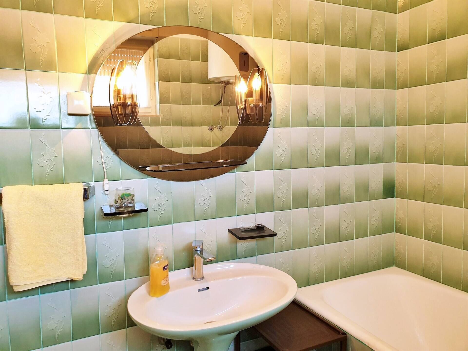 Un bagno con piastrelle verde decorate, uno specchio rotondo, un lavandino, un asciugamano e una bottiglia di sapone.