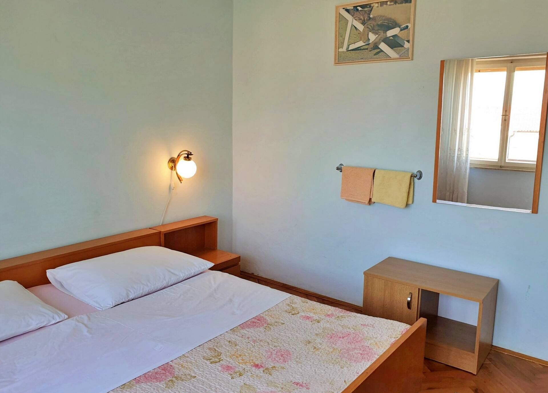A bedroom with a bed, bedside tables, a nightstand, a wall-mounted light, a towel rack, a mirror, and a framed picture.