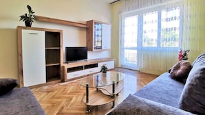 Een woonkamer met een houten visgraatvloer, een glazen salontafel, een flatscreen televisie op een houten standaard, een bank en een potplant.
