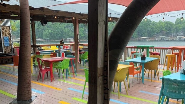 Een terras buiten met kleurrijke stoelen en tafels, houten pilaren en uitzicht op het water.