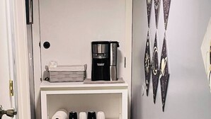 Een hal met een witte console tafel waarop een koffiezetapparaat en kopjes staan, een deur met een greep, en een wand met decoratieve driehoeksmotieven.