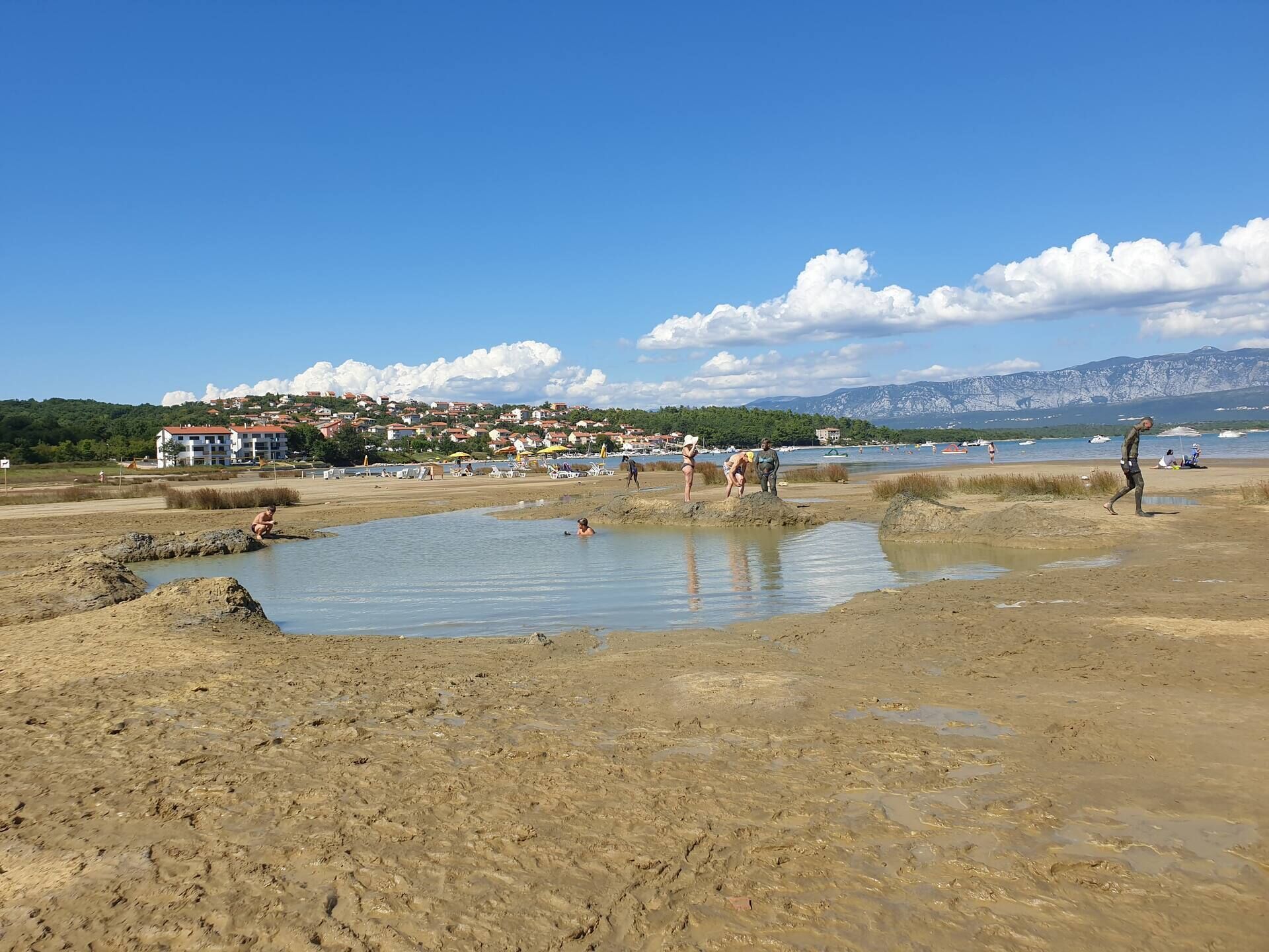 Une plage où l’on pratique diverses activités nautiques, un ciel dégagé et un paysage montagneux en arrière-plan.