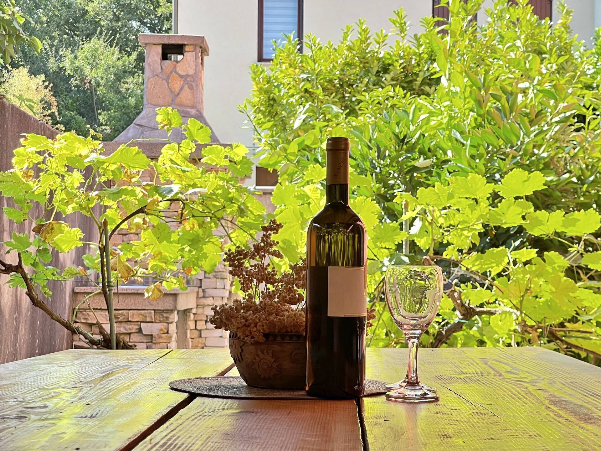 Eine Weinflasche und ein Glas auf einem Holztisch, im Hintergrund ein Steinkamin und üppiges Grün.