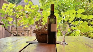 A bottle of wine and a glass on a wooden table with a stone chimney and lush greenery in the background.