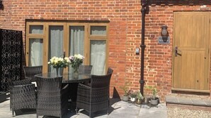Un área de comedor al aire libre con una mesa de vidrio y sillas de mimbre, un muro de ladrillo, una puerta de madera y plantas en macetas.