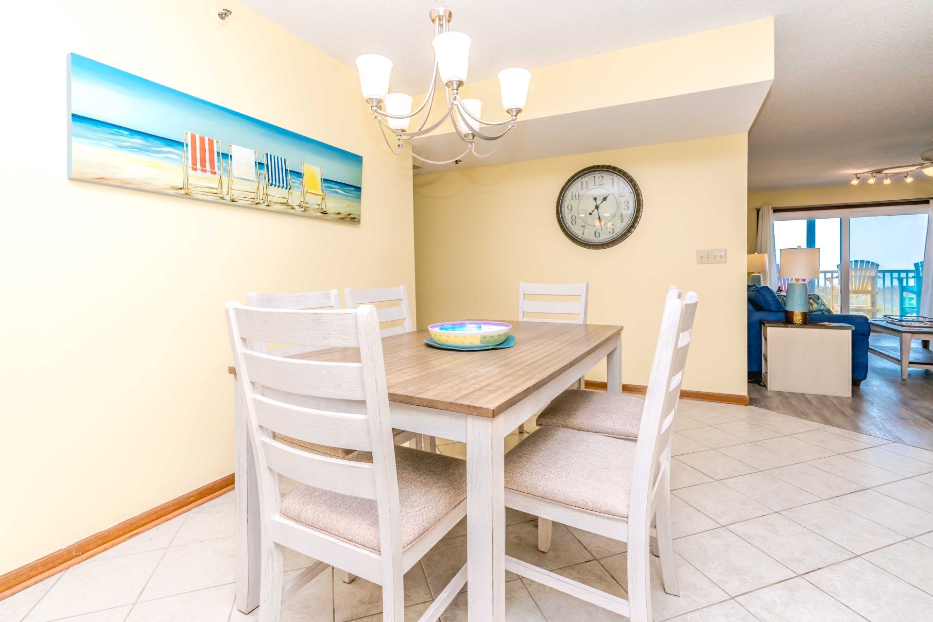 A dining area with a wooden table and white chairs, a painting of a beach scene, a wall clock, and a view of a balcony with a sofa.