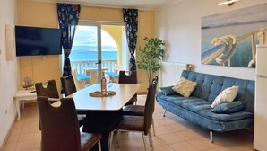 A dining area with a table and chairs, a sofa, a plant, and a view of the sea through a window. - Apartment Neptun - seafront, 20 m from sea, sea view, terrace, private parking (Šilo)