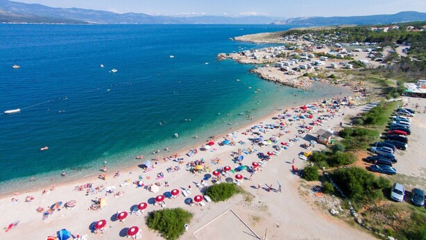 An aerial view of a beach with numerous umbrellas and sun loungers, a parking area with cars, and a clear view of the sea and distant mountains.