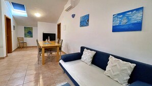 Ein Wohnzimmer mit einer Couch, einem Esstisch und Stühlen. An der Wand hängt ein Gemälde und aus dem Fenster ist der Himmel sichtbar.