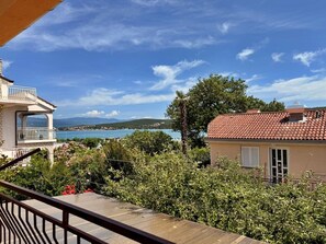 Uma varanda com vista para uma cidade costeira, um edifício com telhado de telhas vermelhas e vegetação exuberante.