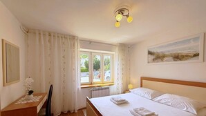 Een slaapkamer met een bed, een bureau, een stoel, een raam met gordijnen, een radiator en een schilderij aan de muur.