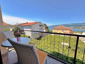 A balcony with a table and chairs, a potted plant, and a view of the sea and buildings.