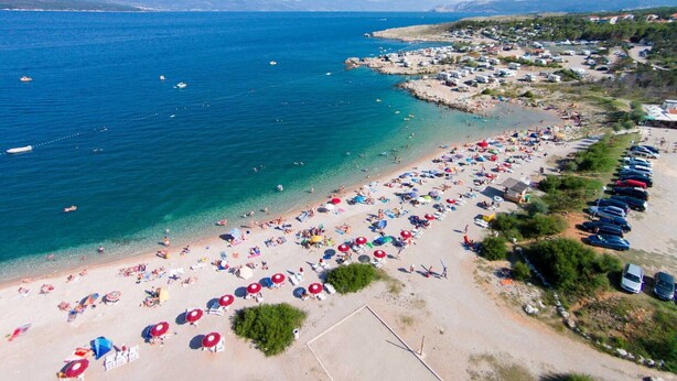 An aerial view of a beach with numerous umbrellas and sun loungers, a parking area with cars, and a clear view of the sea and distant mountains.