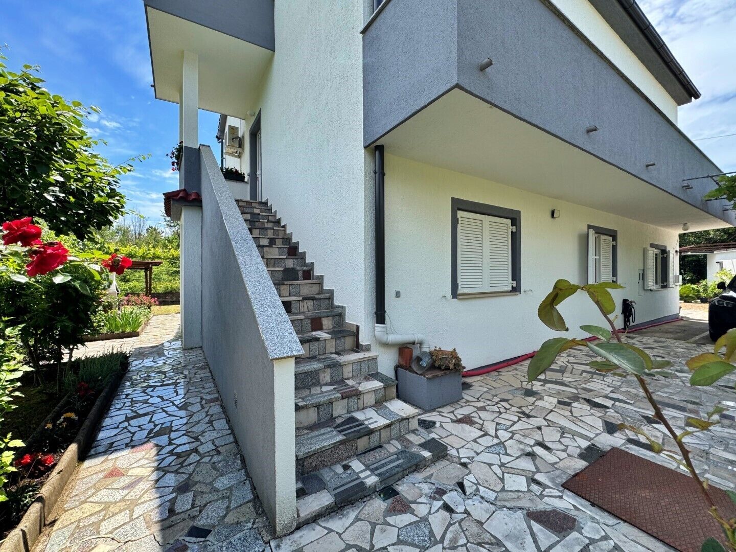 A modern two-story house with a stone staircase leading to the entrance, a covered porch, and a garden with red flowers.