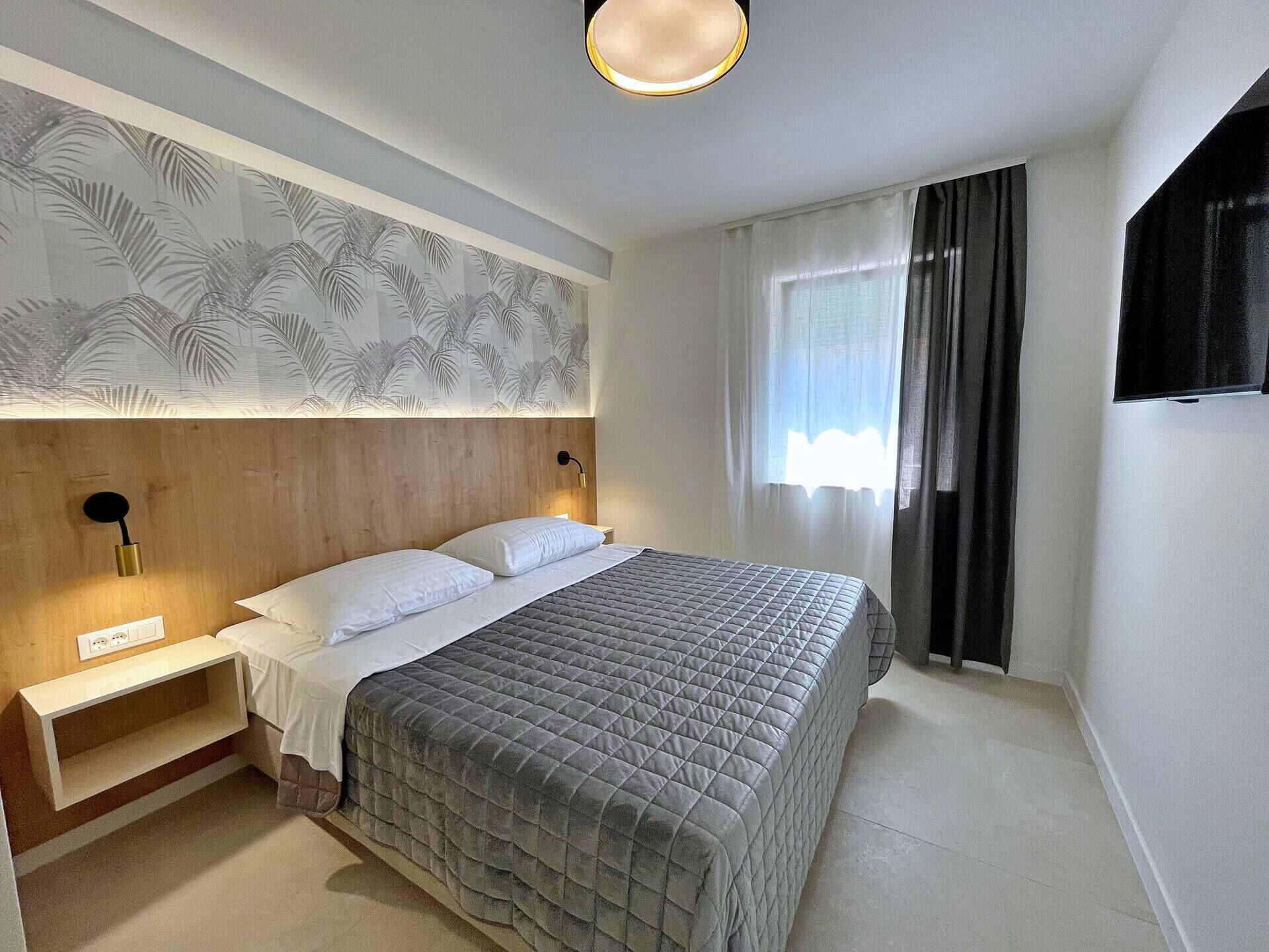 Ett modernt hotellrum med en stor säng, ett sängbord, en väggmonterad lampa och ett fönster med gardiner.