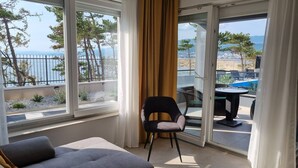 Quarto com uma grande janela que oferece vista para o mar e árvores, uma cadeira e uma mesa pequena.