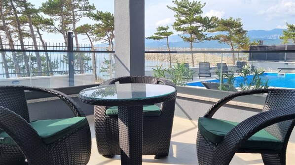 A balcony with wicker furniture, a glass table, and a view of a pool and mountains. - Luxury Apartments Punta Silo - 30 m from the sea, sea view, swimming pool, outdoor shower, Smart TV, laundry room, private parking (Šilo)