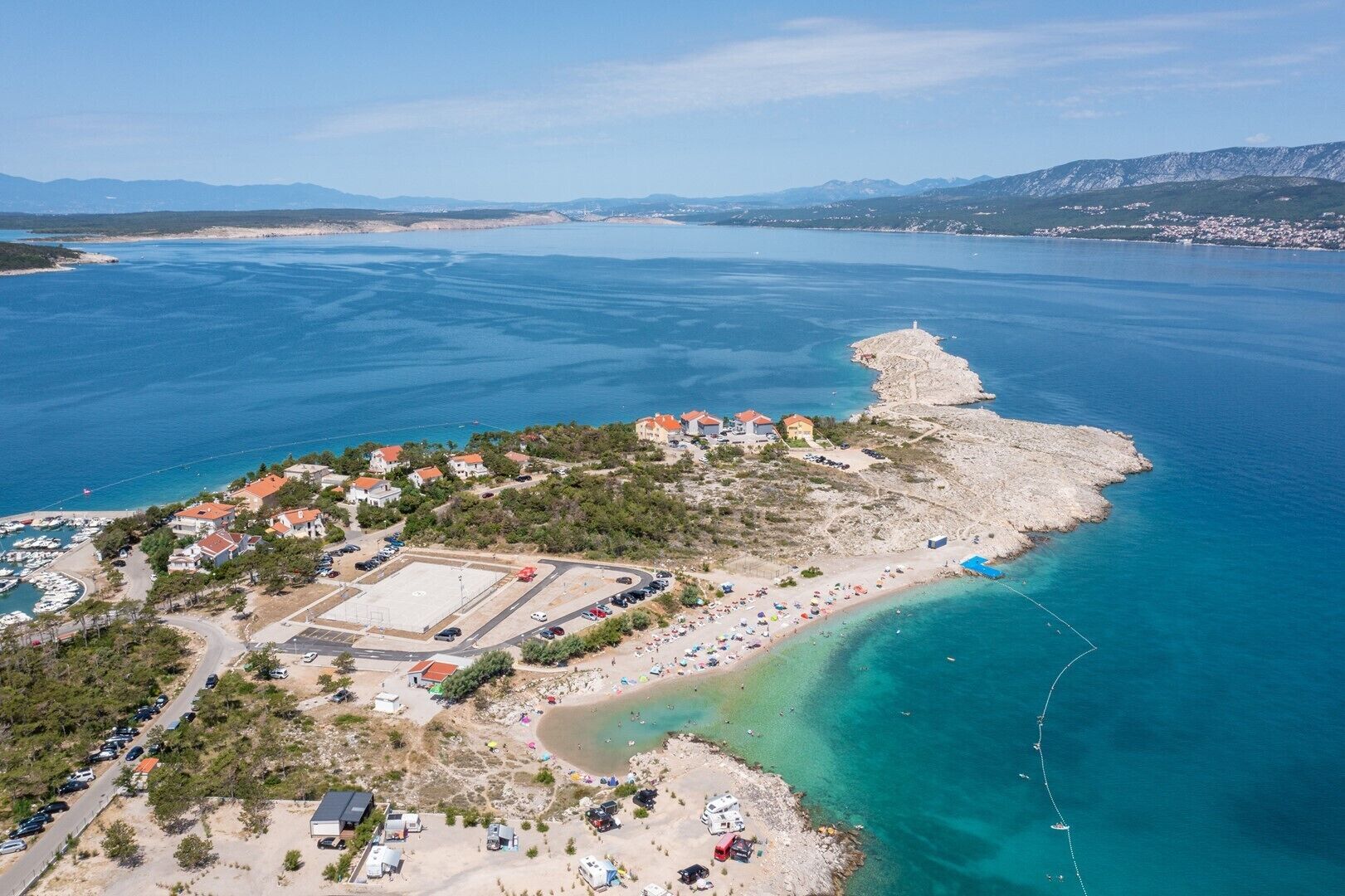 An aerial view of a coastal area with a beach, a marina, and buildings with red roofs.