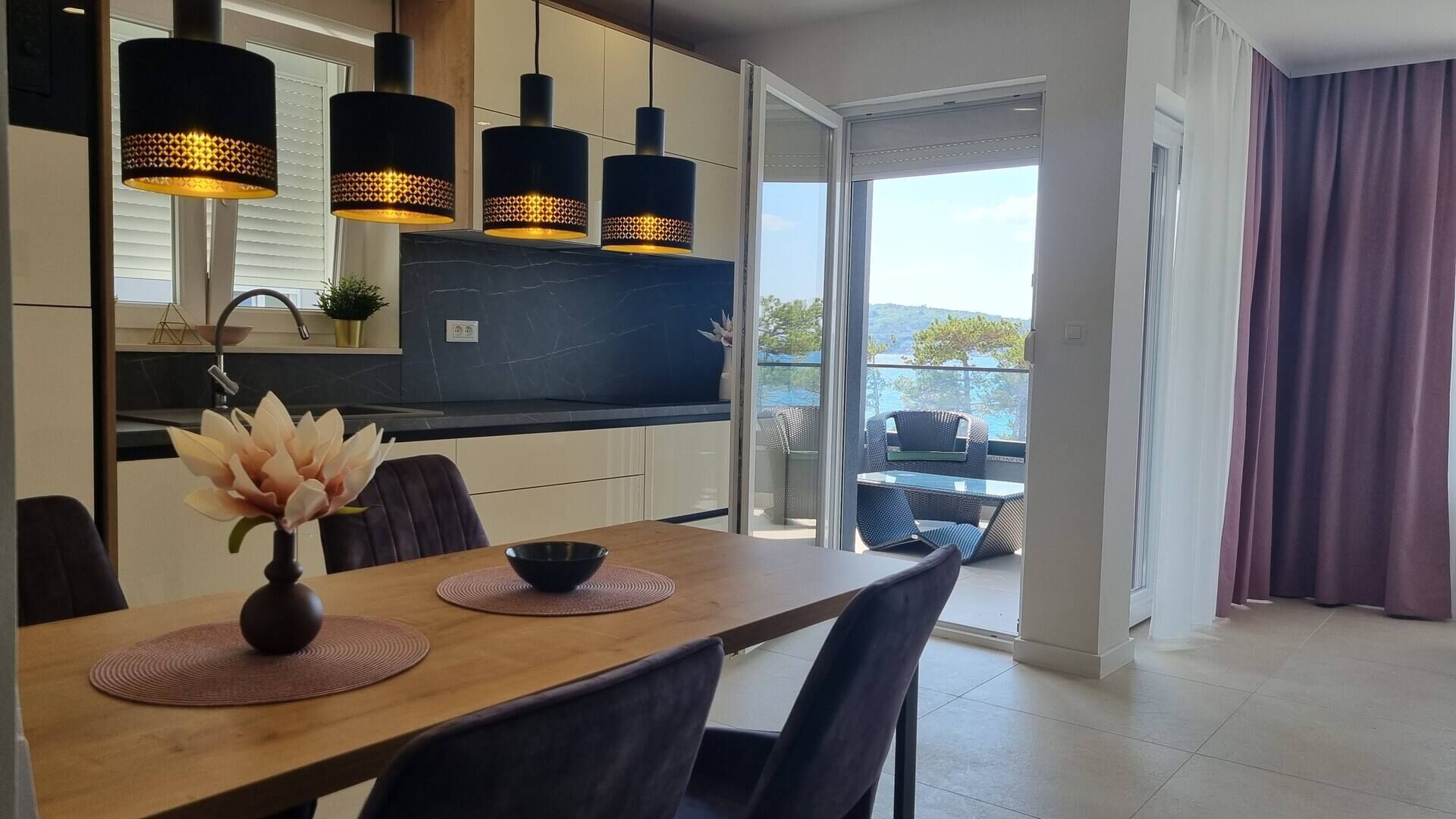 A modern kitchen with dark countertops, a dining area with a wooden table and chairs, and a view of a balcony with outdoor furniture.