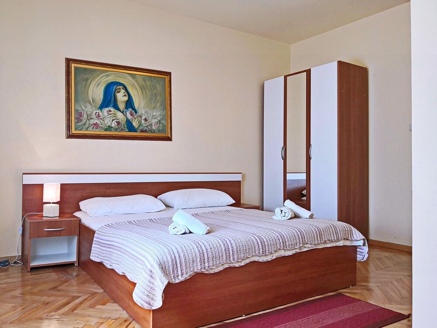 Een slaapkamer met een houten bed, nachtkastje, lamp, kledingkast en een schilderij aan de muur.