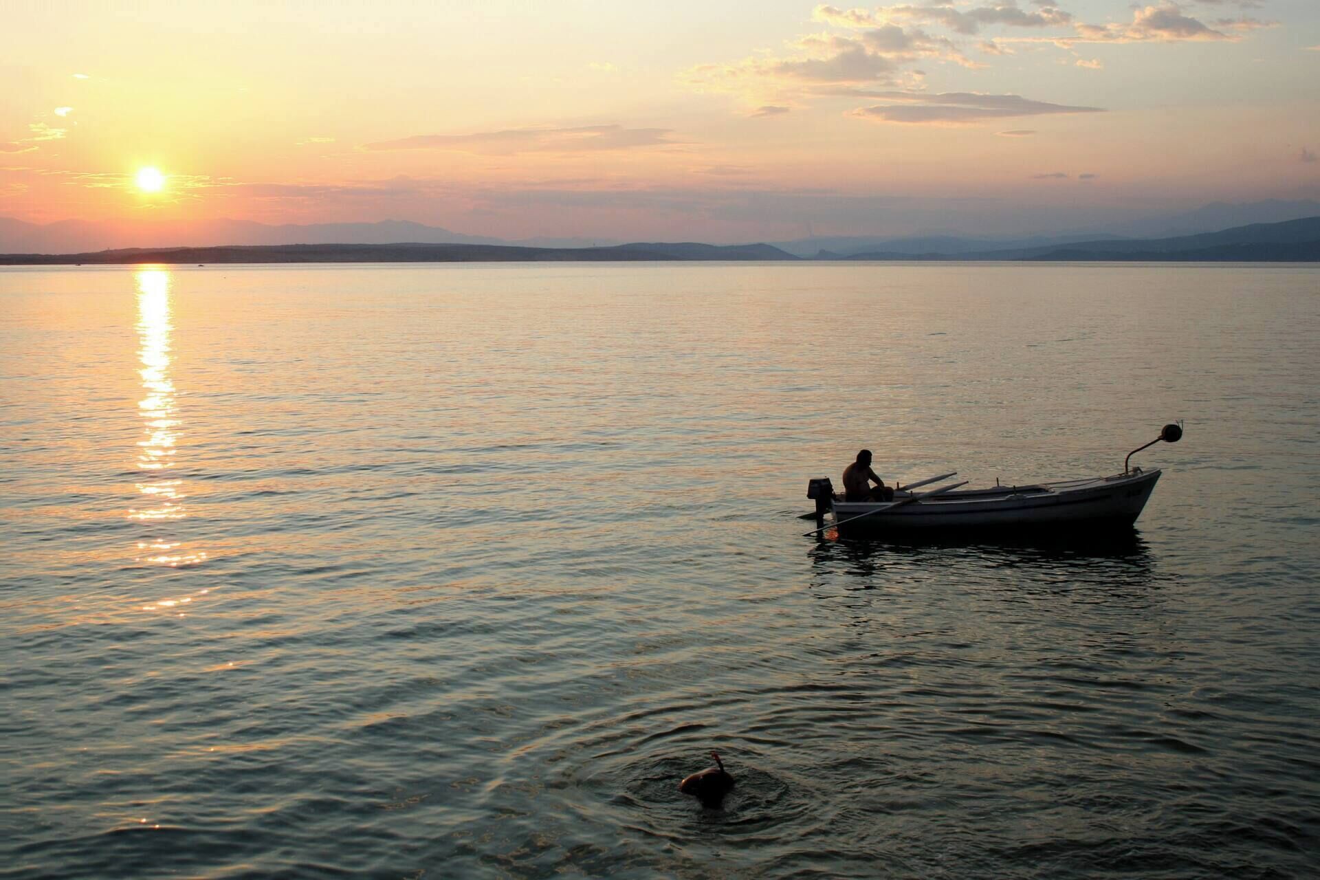 A calm lake at sunset with a boat and a person in the foreground.