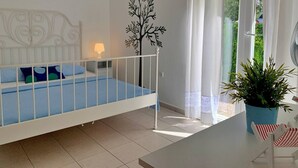 A bedroom with a white bed, a nightstand with a lamp, a wall-mounted tree decal, a window with curtains, and a potted plant on a table. - Apartment Savic - 90 m from the sea, private terrace, air conditioning, private parking, near the sandy beach (Šilo)
