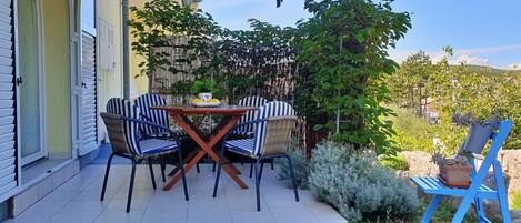 Un patio al aire libre con una mesa, sillas, un paraguas azul y un jardín con plantas y una cerca.