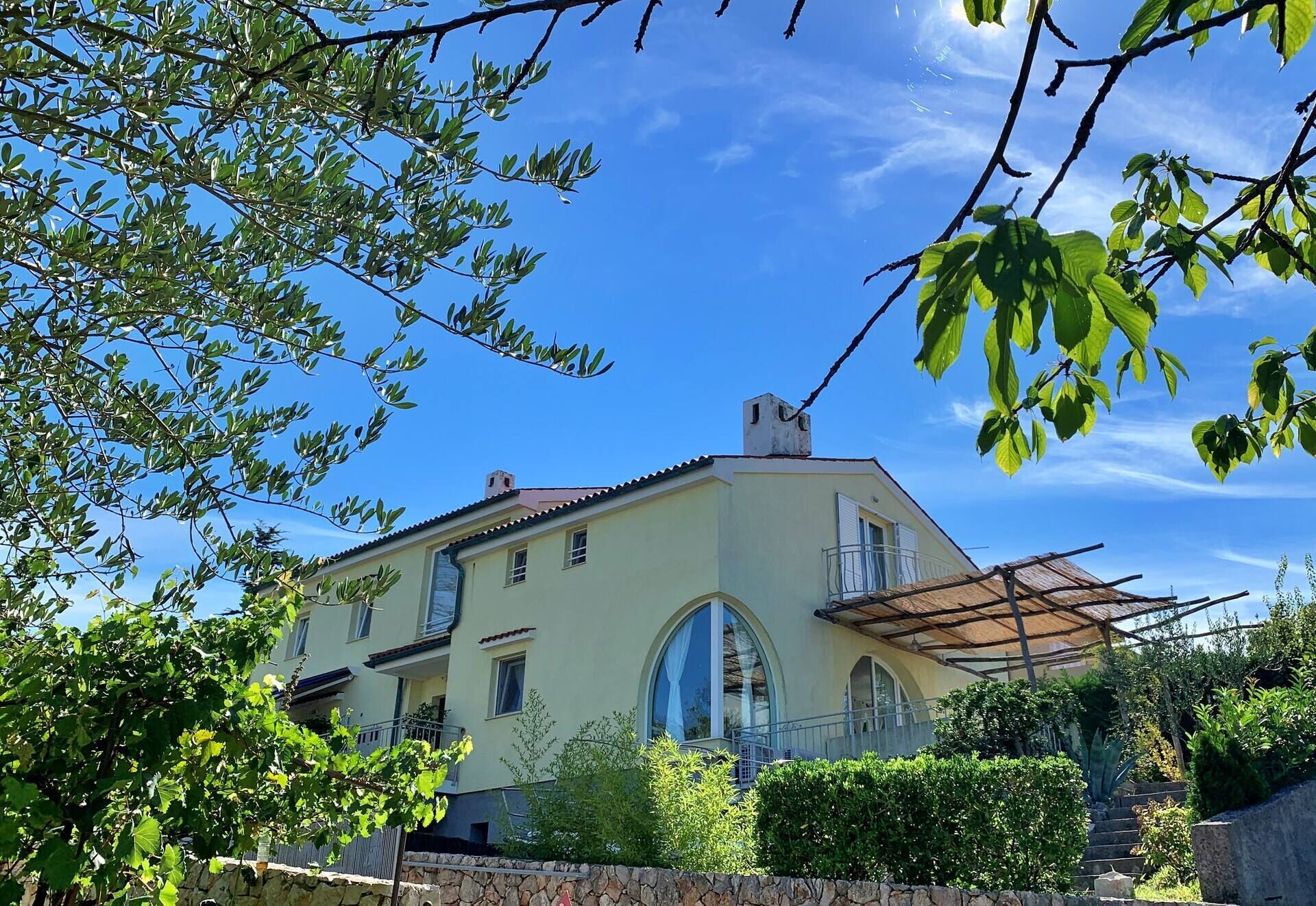 Ein zweistöckiges Gebäude mit hellgelber Fassade, großen Bogenfenstern und einem Balkon mit einer Pergola.