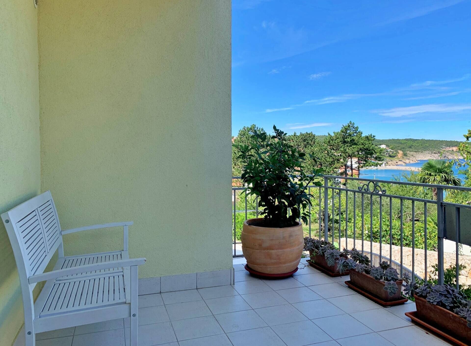 A balcony with a white bench, a large potted plant, and a view of a coastal landscape.