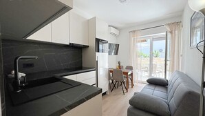 A modern kitchen with black countertops and white cabinets, a dining area with a table and chairs, and a grey sofa in a living area with a large window. - Apartment Scheich - seafront, 20 m from sea, ground floor, terrace (Šilo)