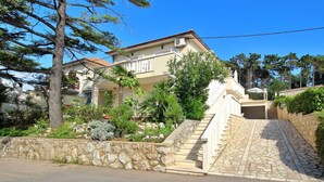 Une maison à deux étages, dotée d’un escalier en pierre menant à l’entrée, et entourée d’une végétation luxuriante et d’arbres.
