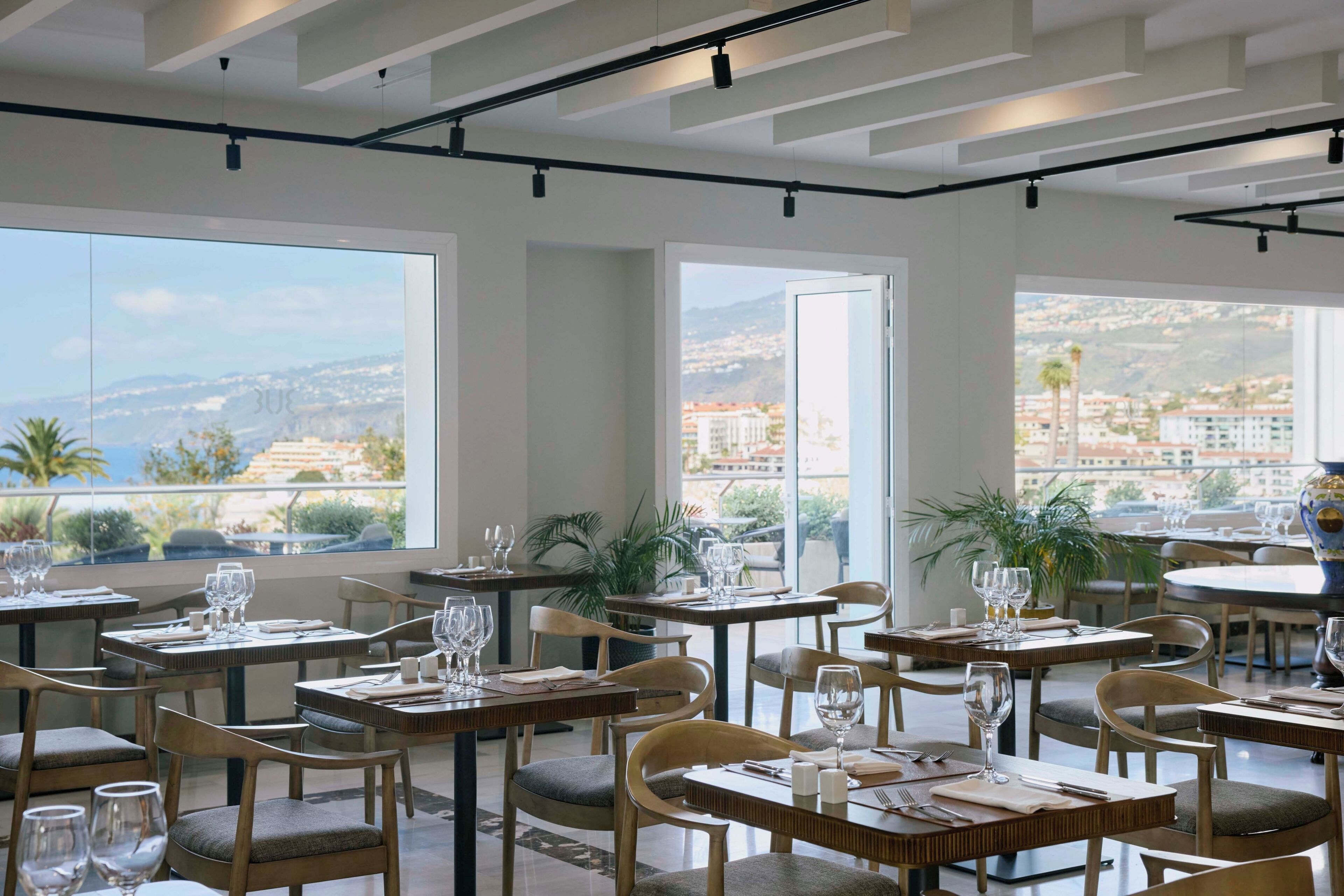 A restaurant with tables set for dining, large windows offering views of the city, and a modern ceiling design.
