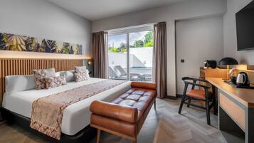 A modern hotel room with a large bed, a bench, a desk with a chair, and a view of the outdoors.