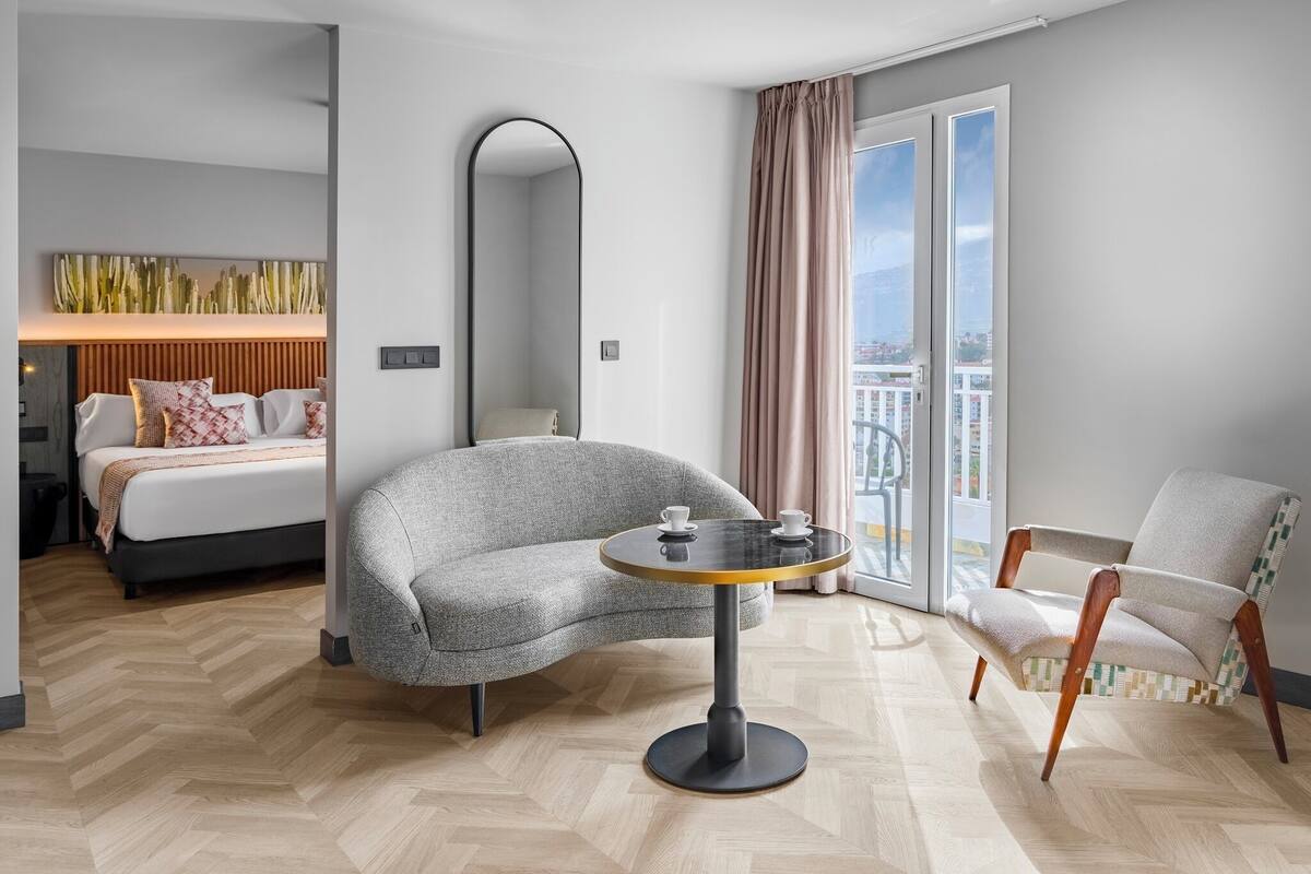 Ein modernes Hotelzimmer mit einem Bett, einem grauen Sessel, einem runden Tisch mit zwei Tassen, einem großen Spiegel und Blick auf die Stadt durch ein Fenster.