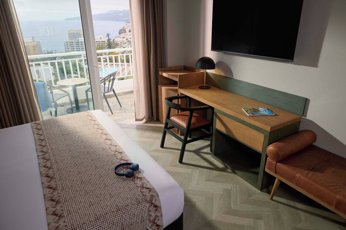 Ein Hotelzimmer mit Bett, Schreibtisch, Sessel, Sofa, Fernseher und Blick auf Stadt und Meer.