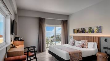 A modern hotel room with a large bed, a desk, a chair, a TV, and a balcony with a view.