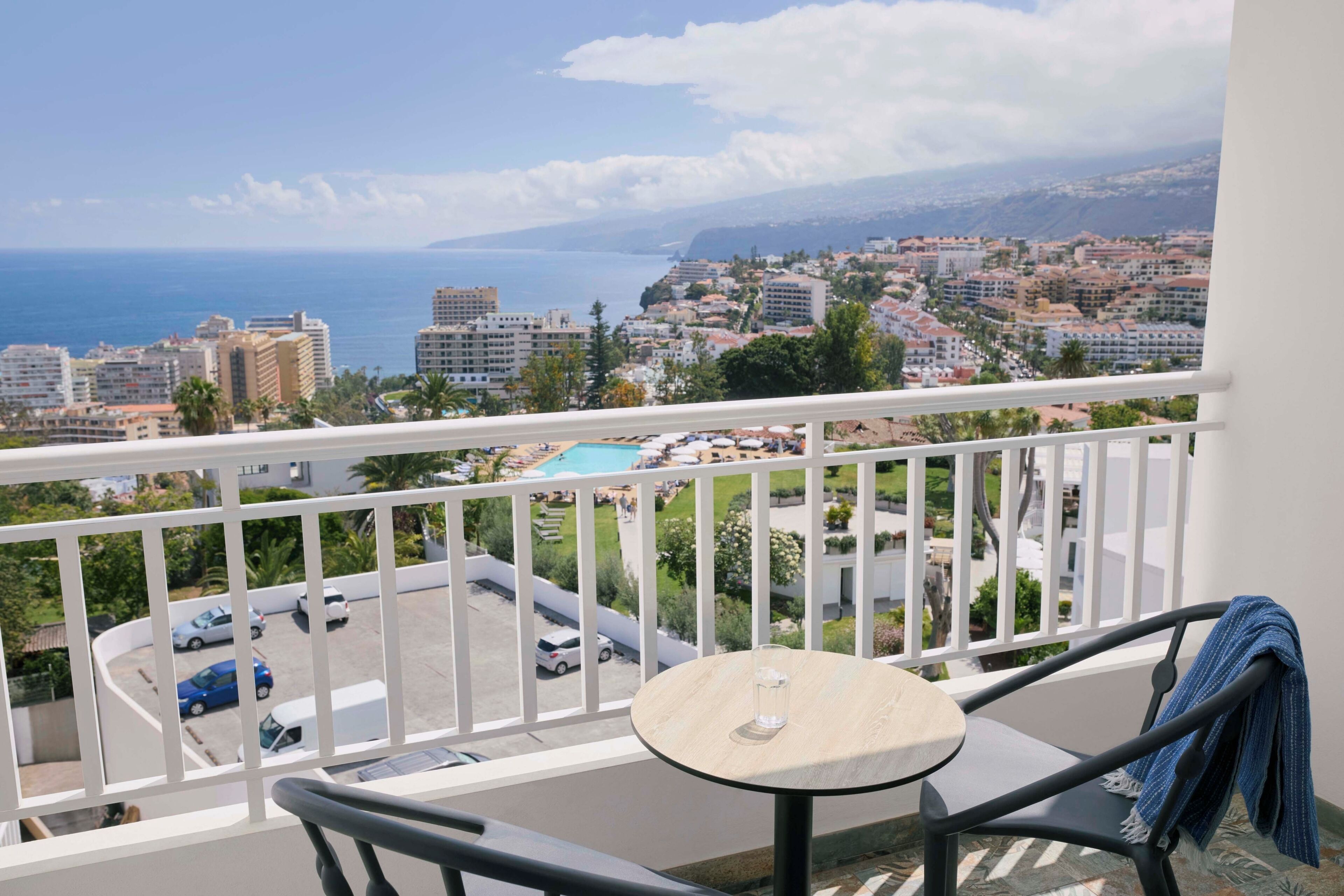 A balcony with a table and chair, overlooking a coastal cityscape with buildings and a parking area.