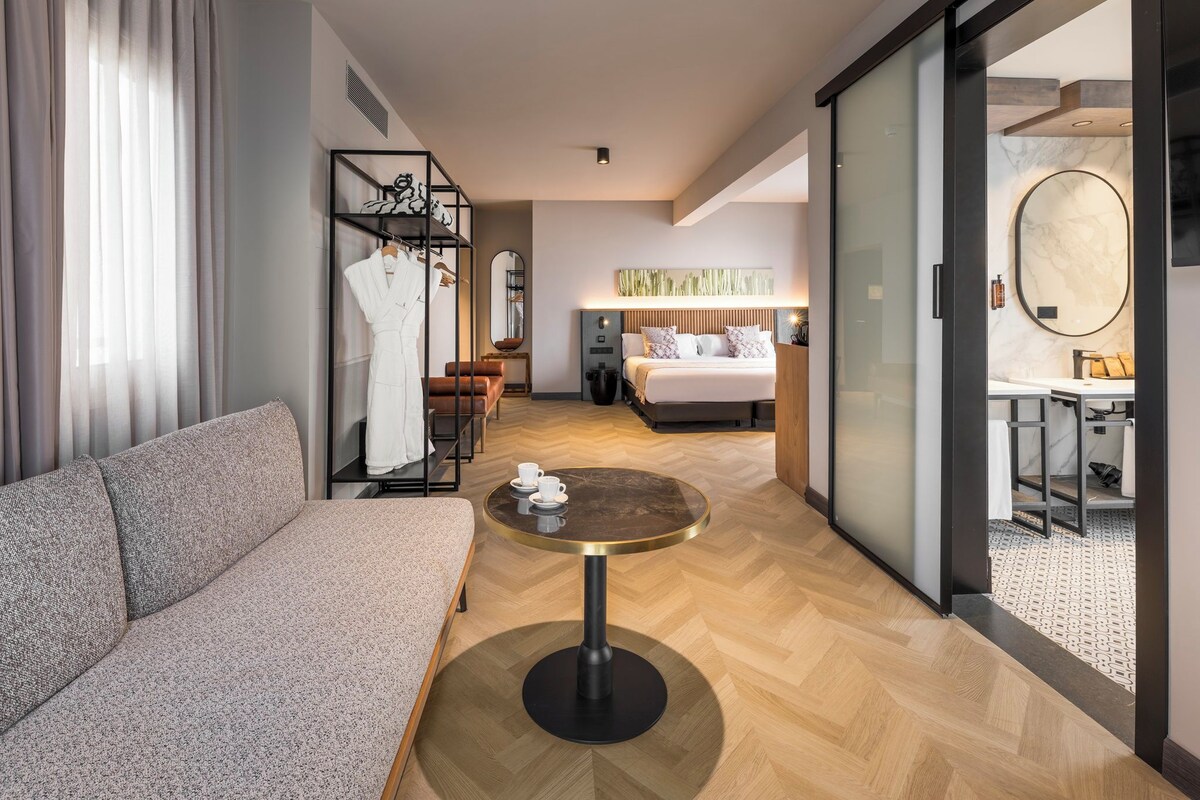 Hotelkamer met een bank, een ronde tafel met een kopje, een bed, een bureau en een spiegel.