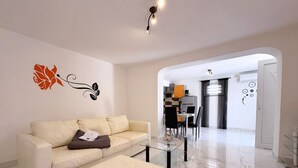 Een moderne woonkamer met een bank, een poef en een glazen salontafel. In de achtergrond is een eethoek met een tafel en stoelen te zien.