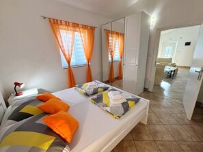Une chambre à coucher avec un lit, des rideaux orange, un bureau avec une lampe, et une salle de bain visible à travers une porte ouverte.